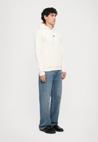 Mann som står med ansiktet mot høyre, iført en off-white hettegenser med frontlomma, blå jeans og svarte sko mot en hvit bakgrunn.