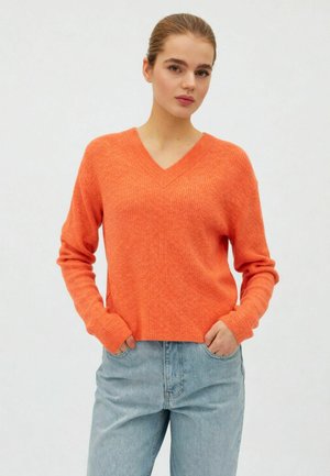 Ung kvinde iført en lys orange V-hals sweater og lyseblå jeans, stående med hænderne i lommerne mod en ensfarvet baggrund.
