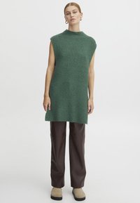 Grüne Strick-Tunika mit hohem Kragen und ärmellosem Design, kombiniert mit braunen Lederhosen und beigen Ankle Boots, getragen von einem Model vor einem schlichten Hintergrund.