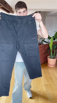 Jauns vīrietis tur melnus cargo bikses ar elastīgu jostu gaišā telpā ar koka grīdu un zaļu augu terakotas podā.