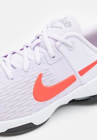 Lyse lilla sportssko med et netoverflade, der har en lys orange Nike swoosh-logo, hvid mellemsål og sort ydersål.