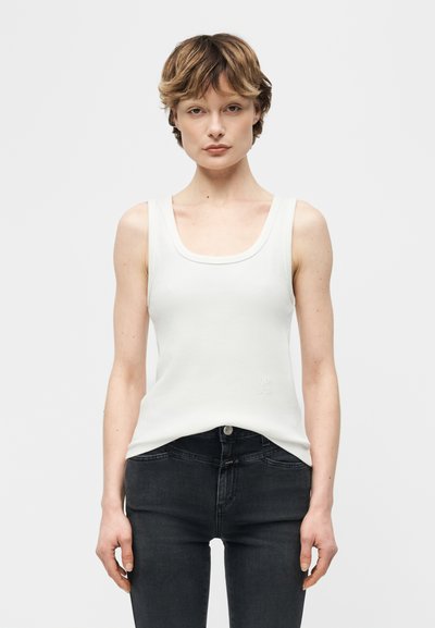 Femme aux cheveux courts portant un débardeur blanc sans manches et un jean noir, debout devant un fond blanc uni.