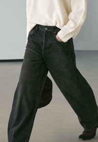 Jeans in denim nero a gamba larga con vita alta, dotati di tasche frontali e chiusura con bottoni, abbinati a un maglione crema dalla texture lavorata.