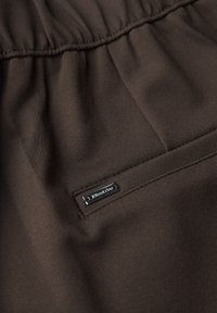 Brun tyg med en slät yta och lätt glans, som har en samlad design längs toppen och en silverfärgad logotag som är sydd.