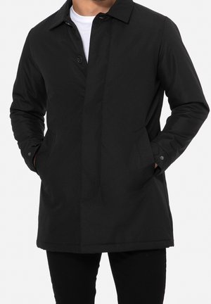 Homme portant un manteau noir boutonné, les mains dans les poches latérales, avec une chemise blanche visible en dessous.