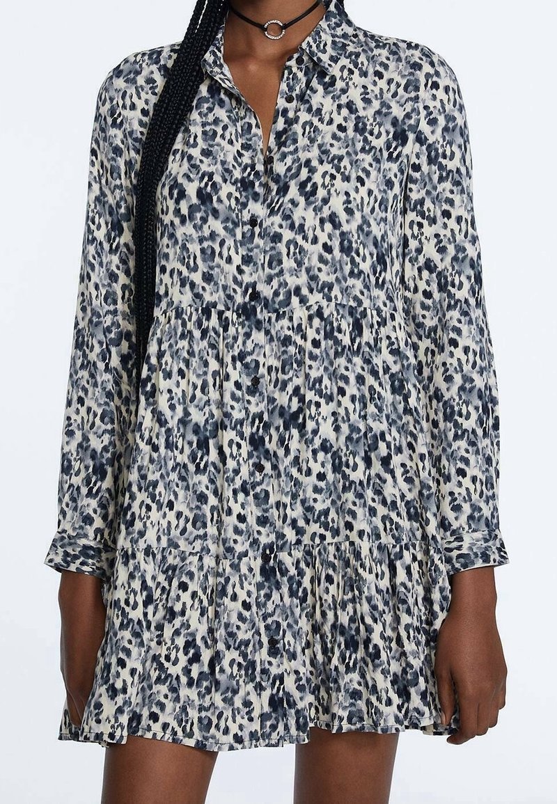 Femme portant une robe à manches longues boutonnée avec un imprimé léopard bleu et blanc et un collier ras-du-cou noir.