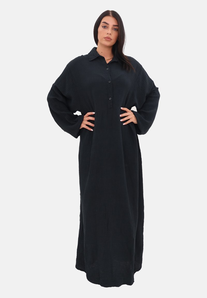 La modella sta in piedi con fiducia, indossando un lungo vestito nero con bottoni e maniche larghe, con le mani sui fianchi, su uno sfondo bianco.