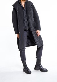 Persoon in een lange zwarte winterjas, een zwart gewatteerd jack, donkere skinny jeans en zwarte stevige leren laarzen tegen een witte achtergrond.