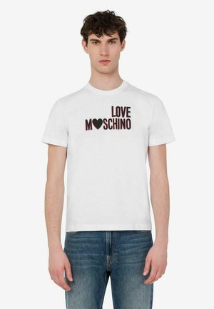 Młody mężczyzna w białej koszulce z napisem "LOVE MOSCHINO" i grafiką serca, w połączeniu z niebieskimi jeansami, stoi na jednolitym tle.