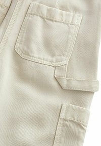 Tessuto beige con una morbida texture, presenta due tasche, una quadrata e una con pattina. I dettagli cuciti aggiungono struttura al design.