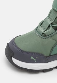 Scarpa sportiva verde e grigia con una tomaia testurizzata, materiali sintetici, punta rinforzata, lacci elastici e una suola bianca con capacità di trazione.