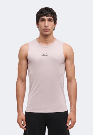 Mężczyzna o krótkich, kręconych włosach, ubrany w jasnoróżową, bezrękawnikową sportową koszulkę Nike Pro oraz czarne spodenki, stojący na jednolitym tle.