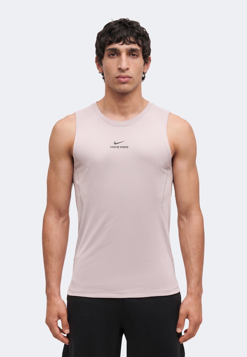 Homme aux cheveux courts et bouclés portant un débardeur de sport Nike Pro rose clair et un short noir, debout devant un fond uni.