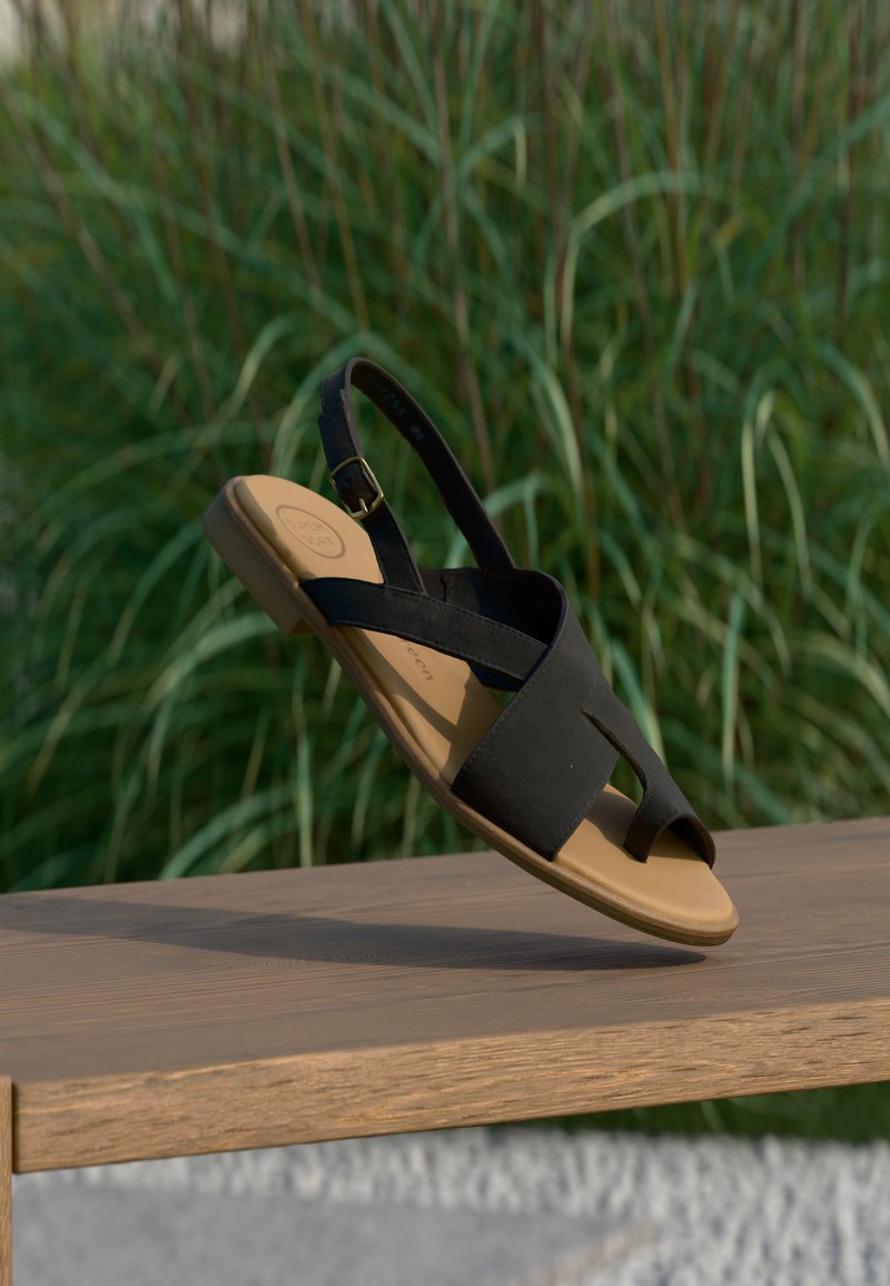 Μαύρο σανδάλι με ταμπά σόλα και λουράκι στον αστράγαλο που ακουμπά σε ξύλινο παγκάκι, με πράσινο ψηλό γρασίδι θαμπωμένο στο φόντο.