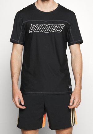 Man die een zwart Adidas T-shirt draagt met een opvallend wit logo en zwarte shorts met beige en oranje verticale strepen.