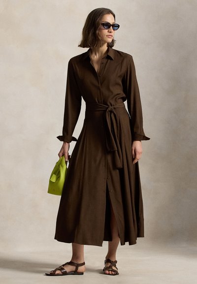 Γυναίκα με καφέ midi φορεμα με μακριά μανίκια και κουμπιά, ζώνη στη μέση, σκούρα γυαλιά ηλίου, σανδάλια και κρατά μια μικρή φωτεινή πράσινη τσάντα.
