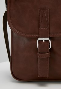 Hnědá kožená taška přes rameno s klopou, stříbrným sponou a nastavitelným popruhem. Hladký povrch a minimalistický design.