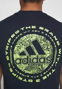 Námořnická modrá bavlněná trička s velkým kulatým grafickým motivem v neonově žluté barvě, na kterém je nápis "adidas" a různé designy související s "třemi pruhy".