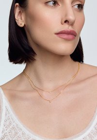 Złote naszyjniki warstwowe z delikatnymi łańcuszkami i małymi, okrągłymi wisiorkami. Modelka nosi przezroczystą, białą bluzkę z koronkowym wzorem.