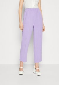 Pantalon taille haute lilas à coupe décontractée, avec une surface texturée et des poches latérales. Associé à des chaussures blanches à talons carrés.