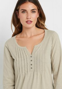 Hellbeige Langarmshirt mit V-Ausschnitt, ausgestattet mit horizontalen Paspeln und fünf Knöpfen an der Knopfleiste. Weicher, strukturierter Stoff.