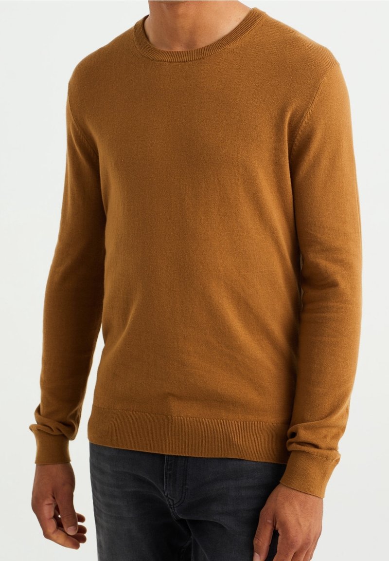 Homme portant un pull à col rond camel à manches longues et un jean foncé, debout devant un fond clair uni.