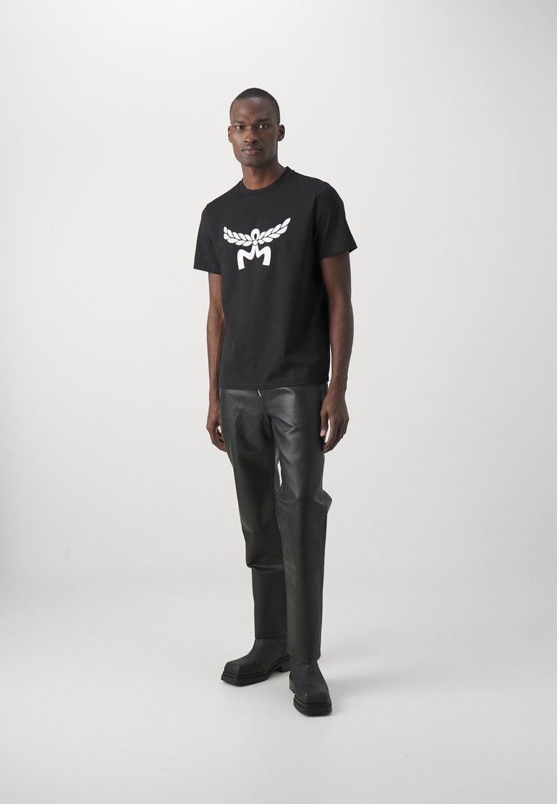 Schwarzes Baumwoll-T-Shirt mit einem weißen Grafikdesign, das einen Lorbeerkranz und den Buchstaben "M" zeigt, kombiniert mit schwarzen Kunstlederhosen und schwarzen Stiefeln.