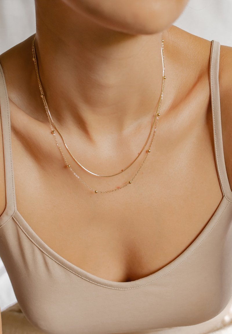 Collane a più strati in tono oro con catene delicate, piccole perline rotonde e una texture liscia e lucida. Indossate sopra un top beige.