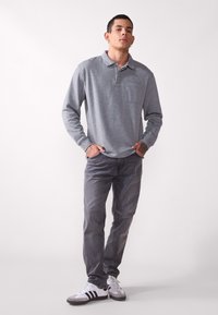 Maglietta polo grigia a maniche lunghe con colletto e tasca anteriore, abbinata a jeans grigi e sneaker bianche con strisce nere. Outfit casual.
