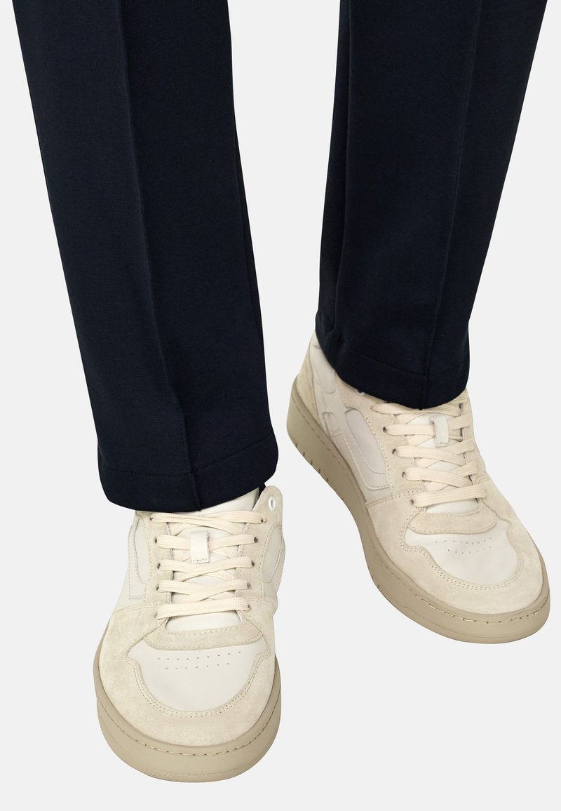 Baskets blanches avec des accents beige portées avec un pantalon bleu marine foncé, montrant les bas de jambes et les pieds sur un fond uni.