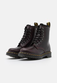 Botas de tornozelo em couro bordeaux com atacadores pretos e costura amarela, apresentando uma sola de borracha texturizada e um forro de pele sintética na gola.