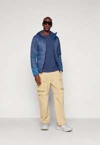 Mėlyna izoliuota striukė su gobtuvu, kreminės spalvos krovinių kelnės, pilki sportbačiai ir tamsūs saulės akiniai; turi dygsniuotą tekstūrą ir šonines kišenes.