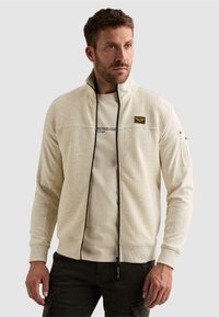 Homme portant une veste zippée texturée beige clair sur un t-shirt graphique et un pantalon foncé sur un fond uni.