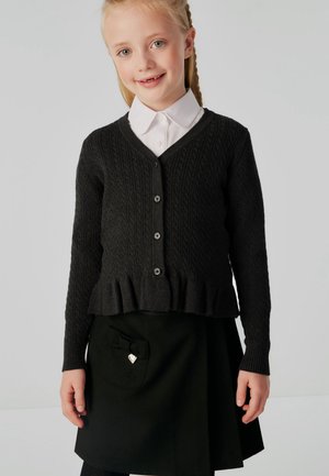 Nuori tyttö, jolla on punotut hiukset, pukeutuu mustaan palmikkoneuleeseen, jossa on napit, valkoisen kauluksellisen paidan päälle ja mustaan hameeseen, jossa on taskun rusettidetalji.