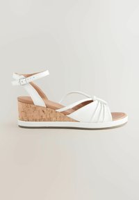 White wedge sandal featuring cork heel, adjustable ankle strap, and twisted front design. Smooth texture with a cushioned sole and contrast stitching.