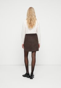 Blonde persoon die met de rug naar ons staat, gekleed in een witte lange mouw shirt, een donkerbruine rok, zwarte doorschijnende panty’s en zwarte hoge hakken.