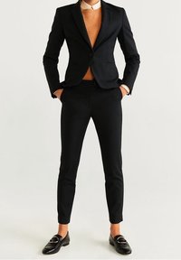 Costume noir sur mesure avec une veste cintrée et un pantalon slim, assorti d'un pull orange et de mocassins noirs. Tissu lisse et design structuré.