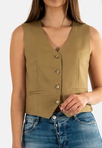 Chaleco de lino en verde oliva con escote en V, seis botones en la parte delantera y dos bolsillos laterales. Lleva puesto sobre unos pantalones de mezclilla azul claro.