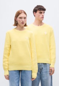 Κίτρινα πλεκτά πουλόβερ με ριγέ μανσέτες και στρίφωμα, με ένα μικρό, διαμαντένιο σχέδιο στο στήθος. Τα μοντέλα φορούν ανοιχτόχρωμο μπλε τζιν.