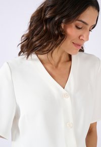 Blusa bianca a maniche corte con scollo a V, caratterizzata da due grandi bottoni rotondi e traslucidi e un tessuto liscio, leggermente strutturato.