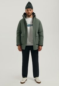 Groene pufferjack met voorzakken, gedragen over een grijze sweatshirt met blauwe tekst, gecombineerd met zwarte broek en witte sneakers.