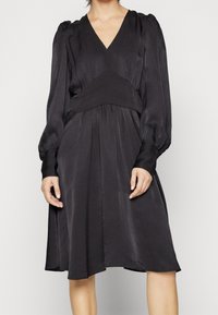 Svart, silkeslen klänning med V-ringning, långa ballongsärmar och en figursydd midja. Faller till en knälång kant och har en mjuk textur och subtil lyster.