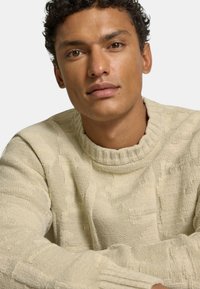 Ljust beige stickad tröja med en hög, ribbad halsringning. Har texturerade mönster. Tyget har ett mjukt och tjockt utseende.