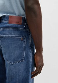 Jeans in denim blu scuro, con una toppa in pelle marrone recante il logo "BOSS" e una tasca con dettagli in bronzo sul retro.