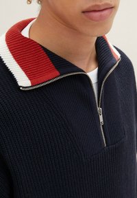 Nahaufnahme einer Person, die einen marineblauen Strickpullover mit einem halben Reißverschluss und einem gerippten Kragen trägt, der rot, weiß und marineblau gestreift ist.