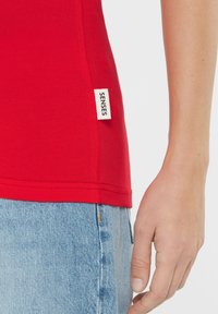 Red cotton t-shirt with a rounded neckline and short sleeves, featuring a small white label with "SENSES" on the side. Denim shorts are partially visible.