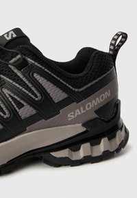 Zwarte en grijze sportieve schoen met een mesh bovenwerk, textuuraccenten, opvallend merk, en een robuuste buitenzool met duidelijke looppatronen.