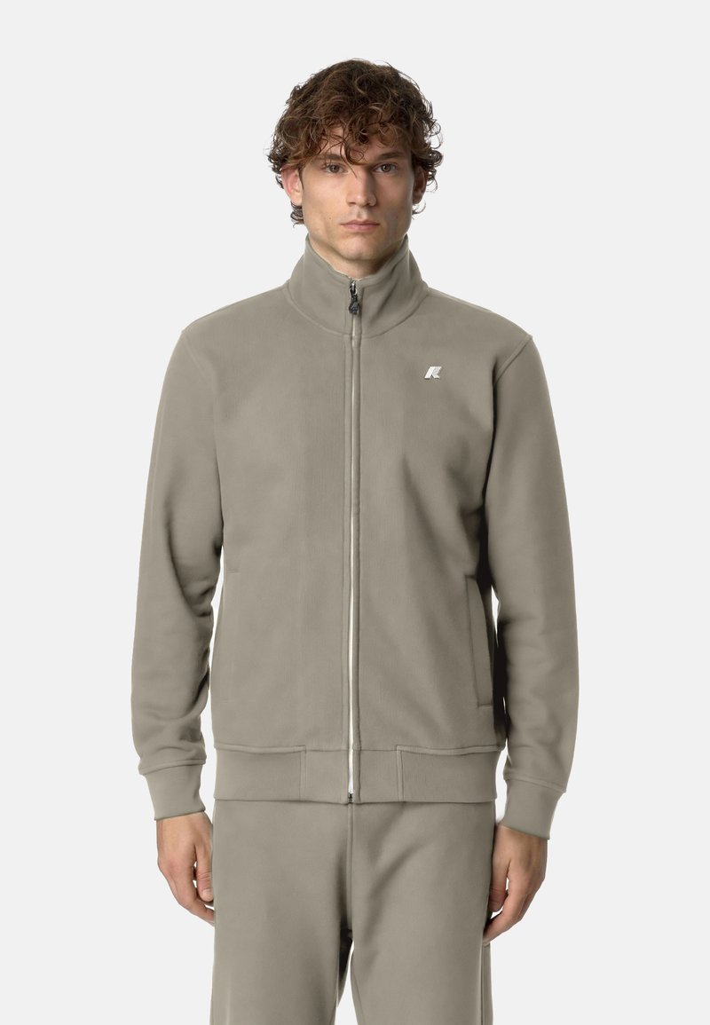 Giacca grigia con zip realizzata in tessuto morbido, con collo alto e due tasche frontali. Presenta un piccolo logo nella zona sinistra del petto.