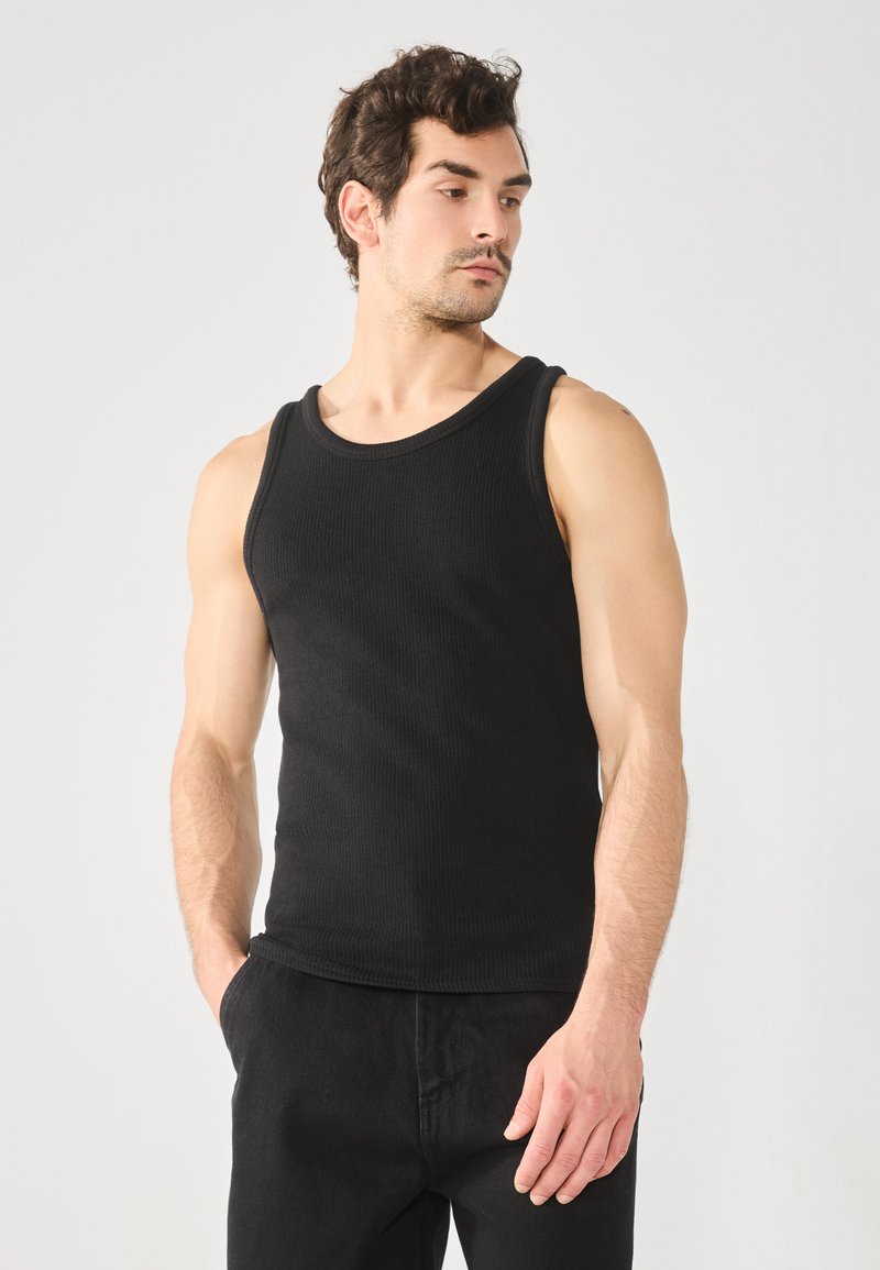 Débardeur noir côtelé avec un col rond et de larges bretelles, porté avec un pantalon noir. Le tissu semble texturé et moulant.