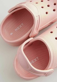 Zuecos de goma rosa con agujeros de ventilación y una correa trasera, que presentan una plantilla texturizada y el logo "NEXT" en relieve en la suela interior.